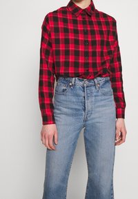 Red and black plaid button-up shirt with a collar, paired with light blue high-waisted jeans. Shirt features black buttons and long sleeves.