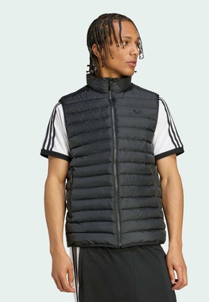 Męski model z warkoczami, ubrany w czarną, pikowaną kamizelkę bez rękawów na białej koszulce Adidas z czarnymi paskami, zwrócony w prawo.