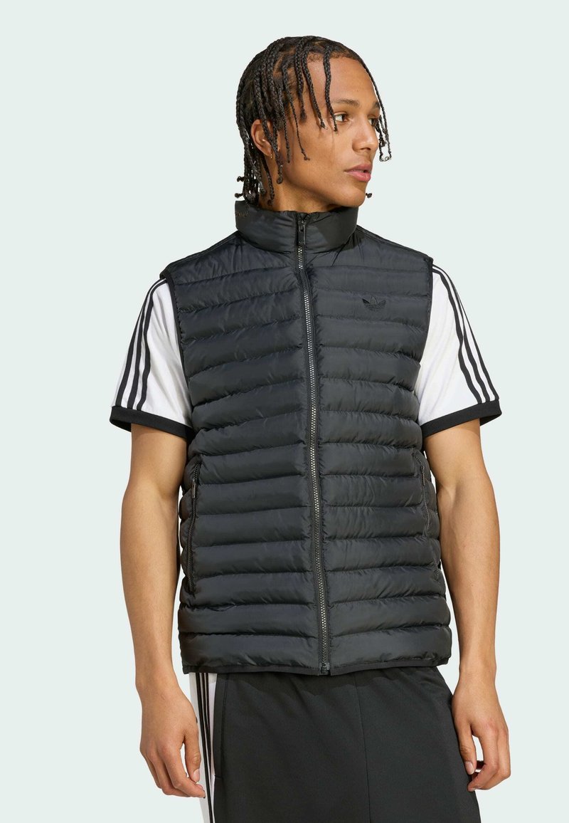 Mandal model med flettet hår iført sort quiltet ærmeløs vest over hvid Adidas skjorte med sorte striber, vendt mod højre.
