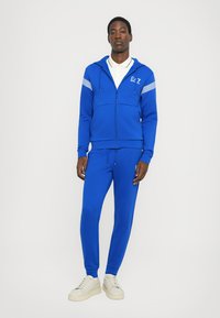 Blue zip-up hoodie with white stripes on shoulders, paired with matching blue sweatpants. Soft fabric, casual design, and fitted silhouette.