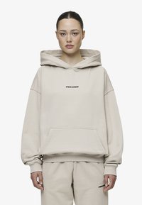 Beige kapucnis pulóver elülső zsebbel, eséses vállakkal és fekete "PEGADOR" logóval a mellrészen. Puha, vastag anyagból készült.