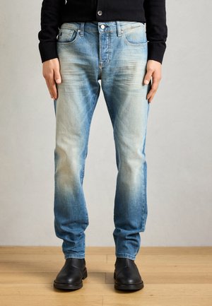 Jean en denim bleu clair avec un motif usé, design à cinq poches et fermeture à bouton en métal. Porté avec des bottes en cuir noir.