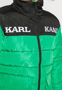 Puchowa kurtka z czarną nylonową górą i jasną zieloną dolną częścią, z białym haftowanym logo "KARL" i zapięciem na zamek. Teksturowana powierzchnia.