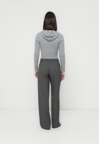 Donna in piedi di spalle, che indossa un maglione grigio aderente con cappuccio e pantaloni grigi larghi, su uno sfondo bianco semplice.