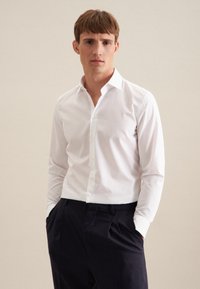 Weißes Langarmhemd mit Button-Down-Kragen aus glattem Stoff. Hat einen figurschmeichelnden Schnitt und wird mit dunklen Hosen getragen, die Hände in den Taschen.