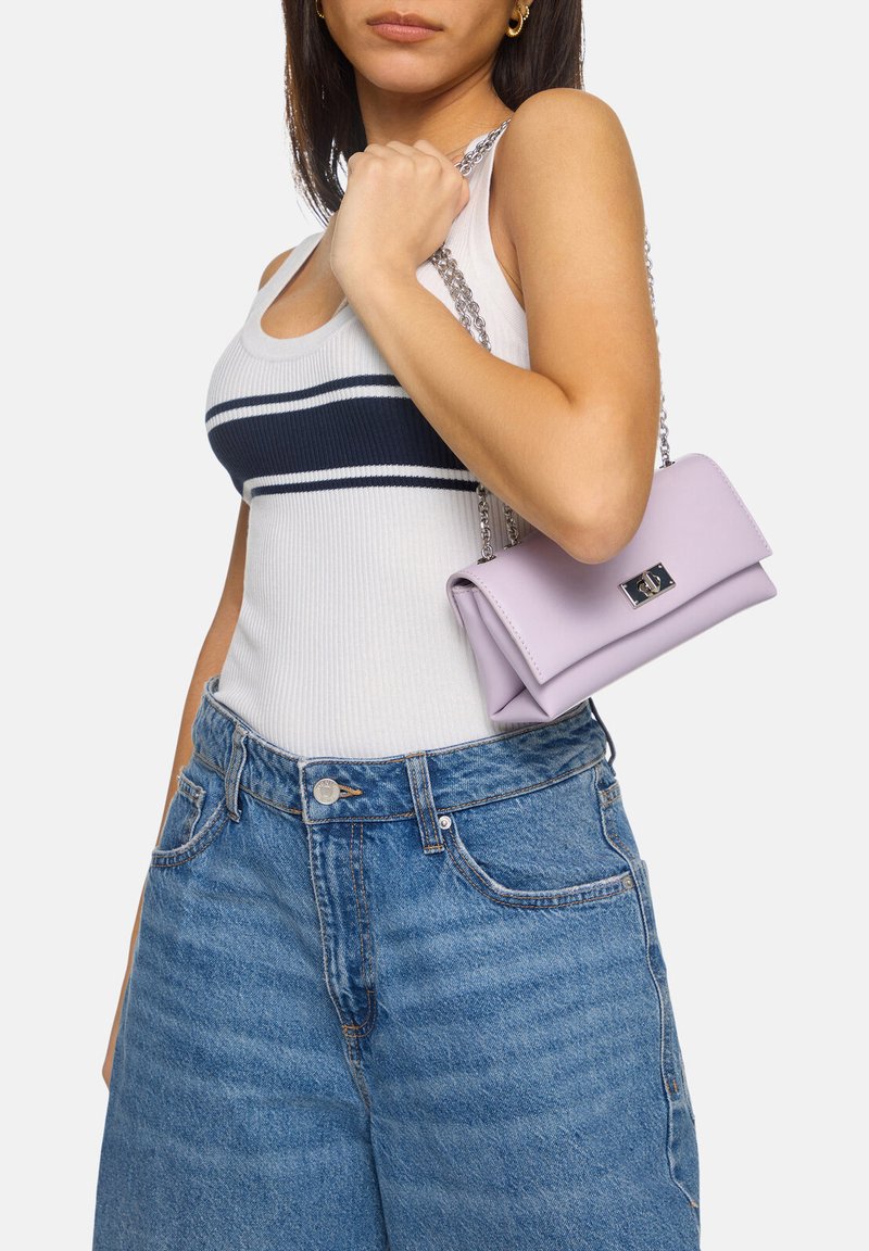 Borsa in pelle lilla con design a sovrapposizione e hardware in argento, dotata di tracolla a catena. Il modello tiene la borsa contro uno sfondo bianco.