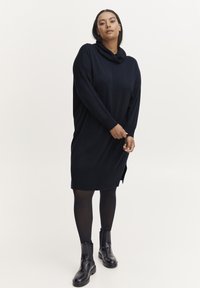 Mornarski oversized pleteni haljinu s kapuljačom, dugim rukavima, bočnim prorezima i rebrastim manžetama, uparenim s crnim čizmama do gležnja.