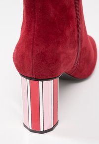 Botte de cheville en daim rouge avec un talon bloc géométrique brillant, présentant des rayures verticales en rouge, rose et blanc. Détail de fermeture éclair sur le côté.