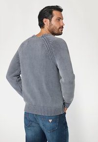 Maglione grigio a maglia con un motivo testurizzato sulle spalle e orlo a coste. Indossato con jeans blu, che mostrano una vestibilità comoda e scollo rotondo.