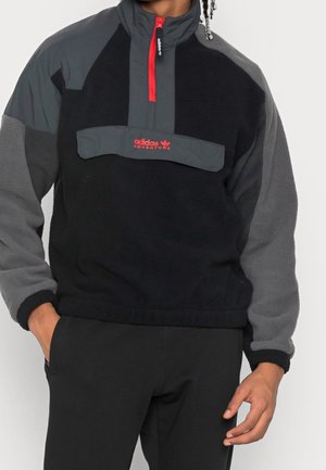 Mand iført sort og grå Adidas fleece-trøje med rød lynlås og logo, hænderne i lommerne på sorte bukser, stående foran en hvid baggrund.