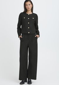 Completo nero a due pezzi con texture, composto da una giacca corta con bottoni dorati e pantaloni a gamba larga, entrambi con tasche laterali.