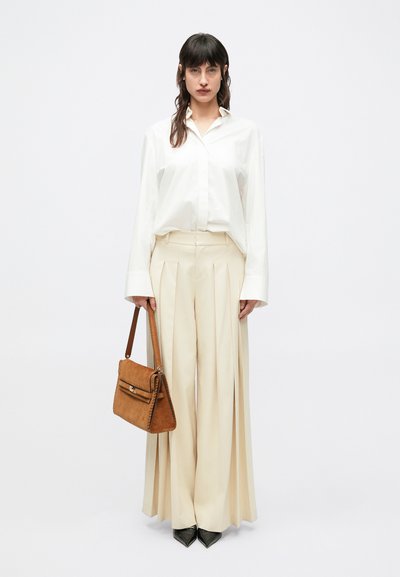 Camicia bianca oversize, pantaloni beige plissettati, tacchi a punta neri e una borsa a tracolla in suede marrone con pattina.