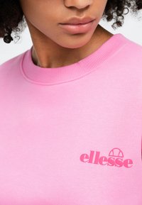 Ružová mikina vyrobená z mäkkého bavlneného materiálu, s rebrovaným výstrihom a malým vyšitým logom "ellesse" na ľavej strane.