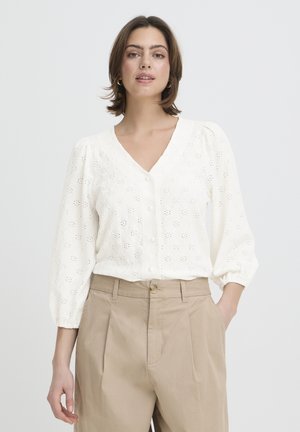 Weiße Lochbluse mit V-Ausschnitt, Knopfverschluss und Puffärmeln. Kombiniert mit hellbeigen, hoch taillierten Hosen mit einer vorderen Falte.