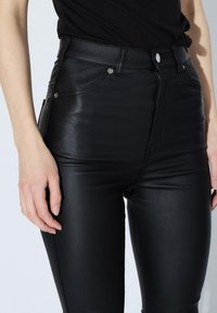 Vysoké černé kalhoty z umělé kůže s hladkým povrchem, předními kapsami a kovovými knoflíky, navržené pro přiléhavý střih.