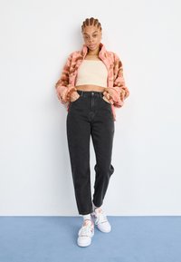 Ružovo-hnedý prúžkovaný chlpatý kabát, béžový crop top, čierne nohavice s vysokým pásom, biele tenisky s farebnými akcentmi. Jednoduché pozadie.