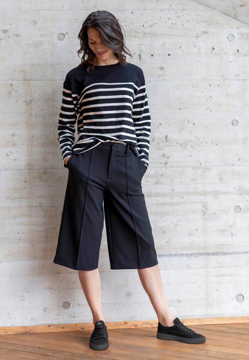 Pasiasta czarna i kremowa koszula z długim rękawem, luźne czarne kuloty, czarne tenisówki; gładkie tekstury i swobodny krój na tle betonu.