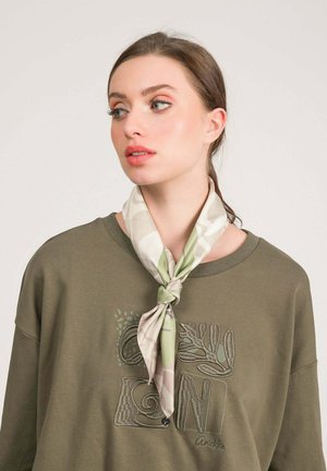 Donna con capelli castani raccolti indossa una felpa verde oliva con un motivo ricamato e una sciarpa leggera a collo con fantasia, annodata sul davanti.