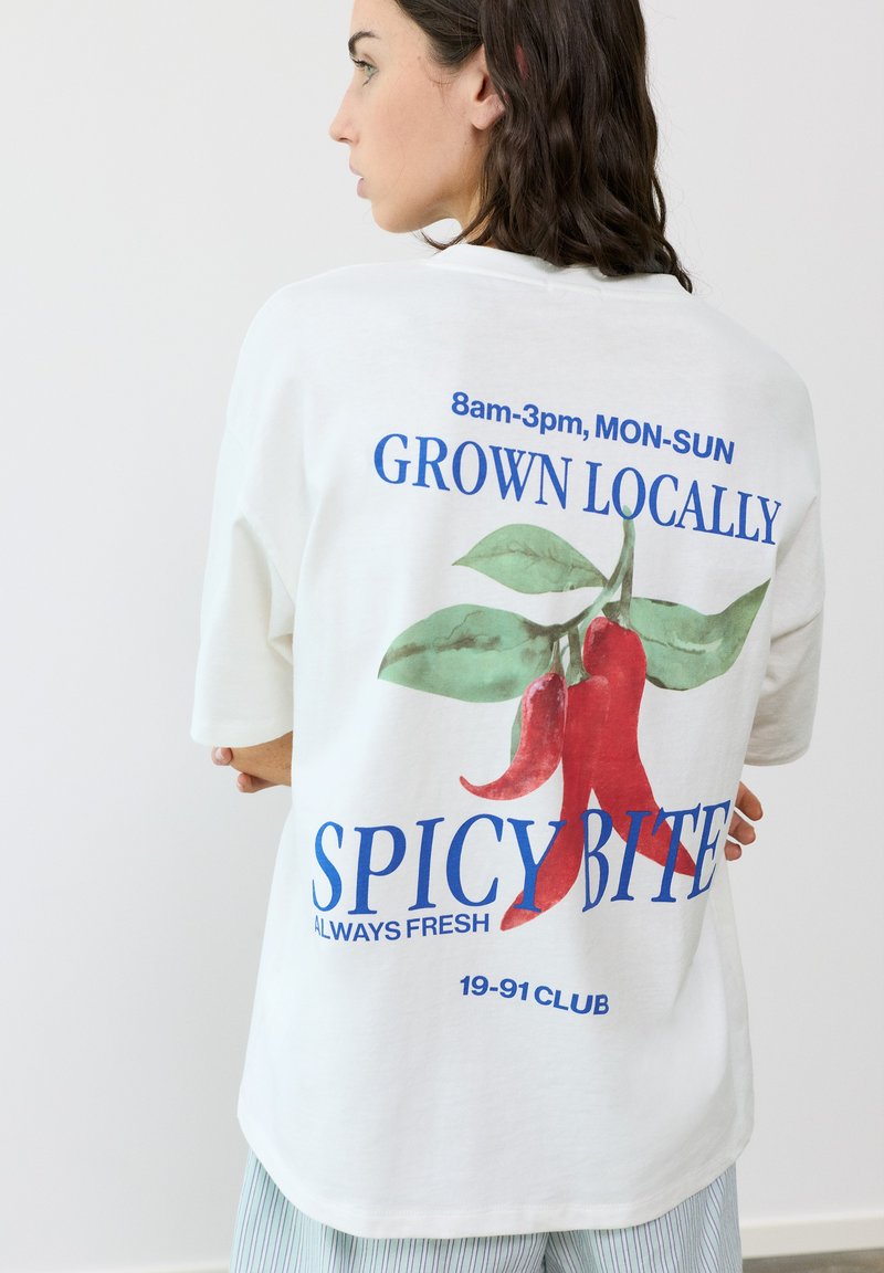 Femme en t-shirt blanc oversize avec des piments rouges et un texte bleu "GROWN LOCALLY SPICY BITE" dans le dos, debout contre un mur blanc.