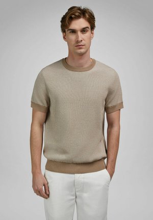 Jeune homme aux cheveux courts et ondulés portant un pull en maille à manches courtes beige et un pantalon blanc, debout devant un fond uni gris clair.