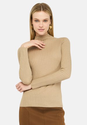 Donna con capelli biondi lisci indossa una maglia beige a maniche lunghe a coste e una gonna marrone, posando con una mano vicino al viso e orecchini a forma di cuore dorati.