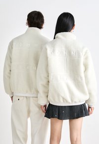 Biele fleezové bundy s textúrovaným povrchom, s logom "TOMMY JEANS" vyrazeným na chrbte.
