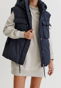Personne portant une robe sweat-shirt gris clair et un gilet matelassé sans manches bleu marine avec de grandes poches avant et une capuche.