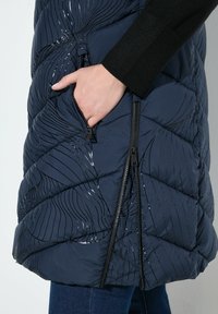 Handen i fickan på en marinblå vadderad jacka med svart abstrakt linjemönster och två synliga svarta dragkedjor, bärs över blå jeans.