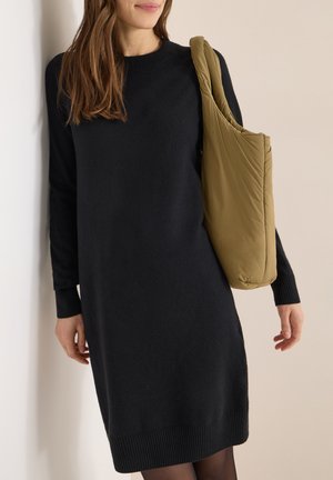 Zwarte gebreide jurk met lange mouwen, geribbelde manchetten en zoom; gecombineerd met een zachte, olijfgroene puffy schoudertas. Over het geheel een gladde textuur.