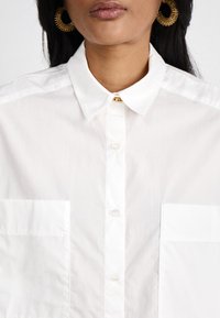 White, collared button-up shirt featuring two chest pockets, smooth texture, and subtle sheen. Accented with a gold detail at the collar.
