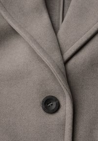 Manteau en laine grise avec une texture douce, présentant des revers notés et un bouton noir gravé. La vue en gros plan met en évidence le détail du tissu.