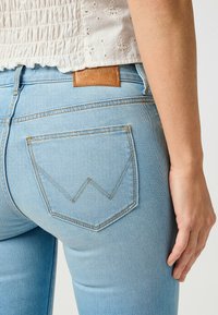 Jean en denim bleu clair avec un design distinctive de poche arrière angulaire, des coutures jaunes et une patch en cuir marron de la marque sur la taille.