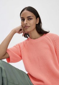 Maglione rosa lavorato a maglia con maniche corte e una texture a coste, abbinato a pantaloni verde oliva. Lo sfondo è bianco semplice.