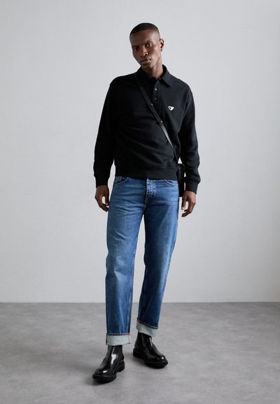 Black long-sleeve polo with a small logo, blue straight-leg jeans, and black ankle boots. Smooth texture and rolled cuffs on jeans.