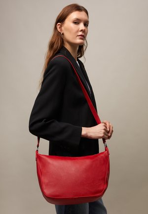 Vrouw met lang bruin haar draagt een zwart colbert en heeft een grote rode leren schoudertas tegen een effen beige achtergrond.