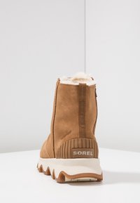 Brązowy zamszowy botek za kostkę z futerkiem, teksturowaną podeszwą i prążkowanym obcasem. Posiada pętelkę do ściągania i tłoczony napis "WODOODPORNE".