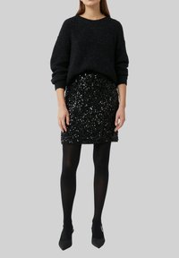 Schwarzer Strickpullover mit lockerer Passform, kombiniert mit einem enganliegenden schwarzen Paillettenrock. Model trägt schwarze Strumpfhosen und spitze Absatzschuhe.