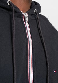 Felpa nera con zip, dotata di una cerniera argentata, con accenti a strisce rosse, bianche e blu lungo la zip. Tessuto strutturato, con cordini regolabili.