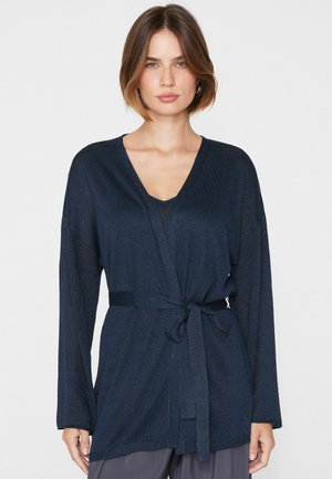 Donna con un cardigan blu scuro a maniche lunghe con cintura annodata in vita, in piedi davanti a uno sfondo chiaro e semplice.