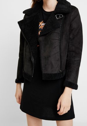 Zwarte faux suède jas met een zwarte fleece kraag en manchetten, voorzien van een diagonale rits en gedetailleerde stiksels bij de schouders.