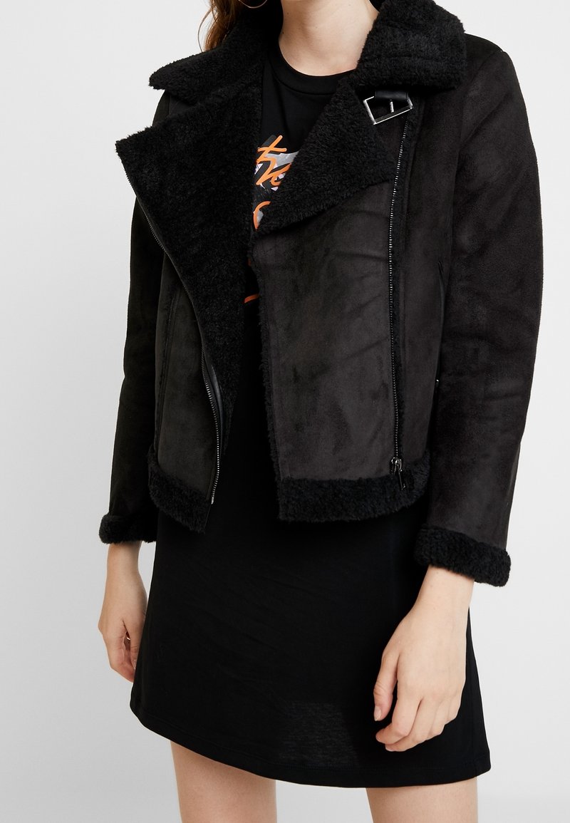 Veste en fausse suède noire avec un col et des poignets en fourrure noire, dotée d'une fermeture éclair diagonale et de coutures détaillées près des épaules.