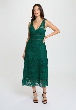 Frau trägt ein ärmelloses grünes Spitzen-Midikleid mit V-Ausschnitt, kombiniert mit schwarzen Riemchen-High Heels, goldenem Armband, Creolen und Halskette.