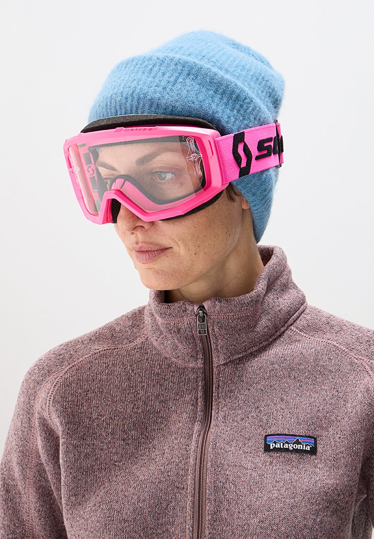 Różowe gogle narciarskie z przezroczystymi soczewkami, czarnym regulowanym paskiem i niebieską dzianinową czapką. Osoba nosi brązową polarową kurtkę na zamek.