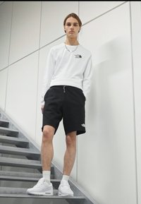 Chemise blanche à manches longues avec logo, shorts noirs et baskets blanches. Se tient sur un escalier avec des marches en métal et un fond minimaliste.