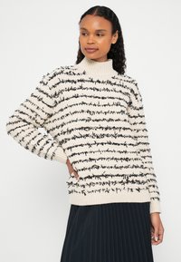 Femme portant un pull à col roulé crème avec des rayures horizontales texturées noires et une jupe plissée noire, posant avec une main sur la hanche.