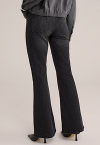Pantalones vaqueros negros acampanados con bolsillos traseros y un acabado de denim texturizado. Un modelo los lleva con tacones de punta para añadir contraste.
