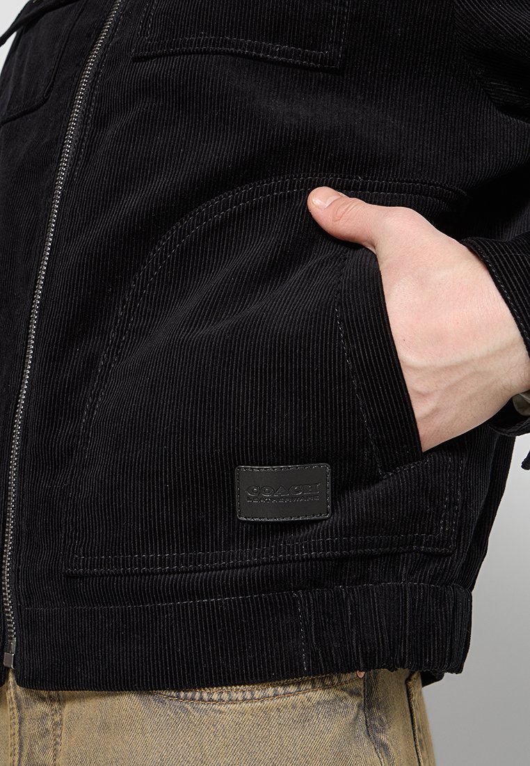 Veste en velours côtelé noir avec fermeture éclair à l'avant, poches latérales, poignets côtelés et motif texturé ; comprend une petite étiquette de marque en cuir rectangulaire.