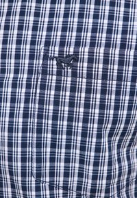 Koszula z krótkim rękawem w granatowo-białą kratę, z kieszenią na lewej piersi z haftowanym logo. Materiał bawełniany, gładka tekstura.