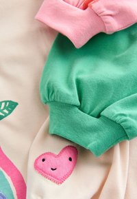 Dos camisas mostradas juntas: una de manga larga verde con puños fruncidos y un puño rosa; una tela ligera con un parche en forma de corazón rosa en la manga.