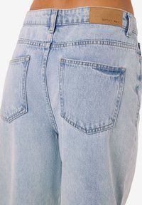 Šviesiai mėlynos spalvos džinsai su laisvu siluetu, turi dvi kišenes nugaroje ir smėlio spalvos etiketę su reljefiniu užrašu „NOISY MAY“ ant juosmens.